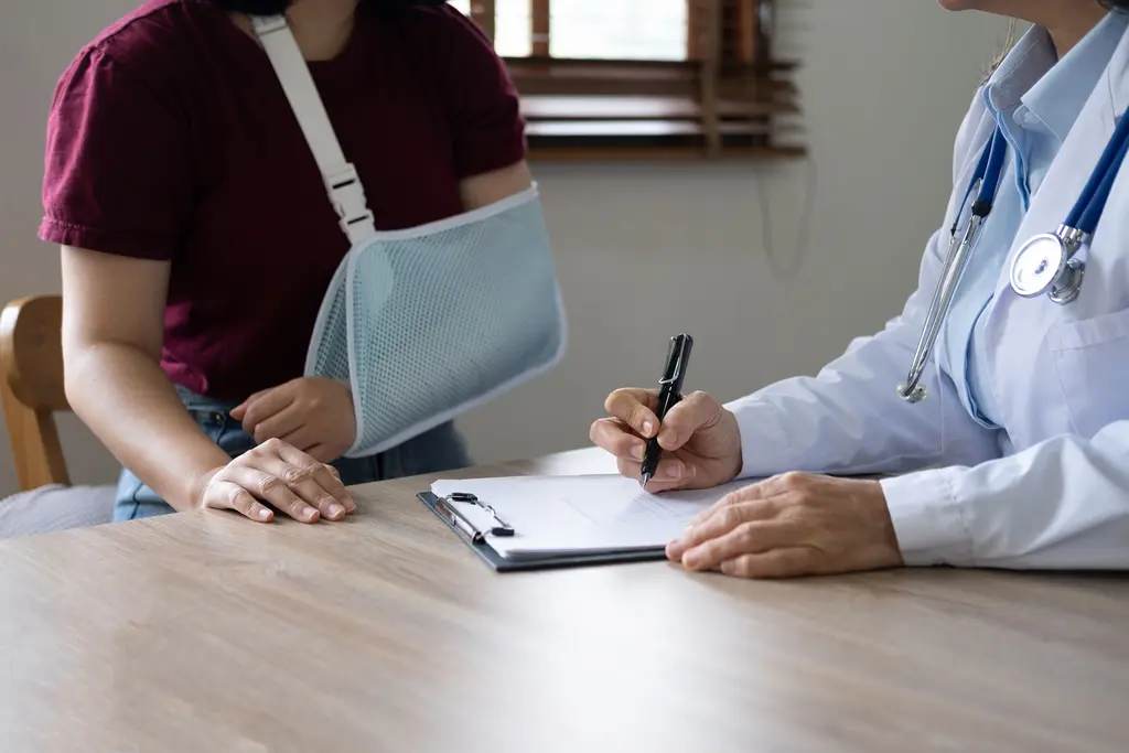 Injured patient wearing an arm sling while a doctor writes notes on a clipboard during a medical consultation.